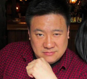 A headshot of improviser Jason Chin. He's weasring a red shirt, looking directly at the camera with his hand on his chin.