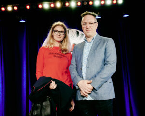 Brunswick partner, Sarah West & AndAlso director, Jules Munns stand on stage in front of the comedy store logo at the finale of a 300 person Christmas team building event.