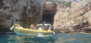 A yellow sea safari boat with guests on, bobs about in a choppy cove with a cave in the background.