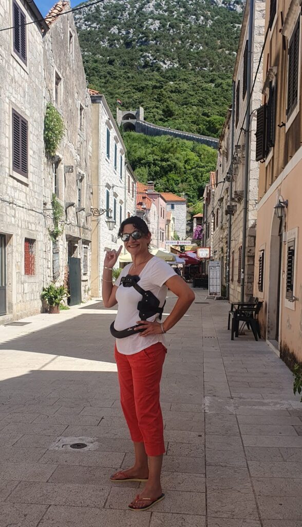 AndAlso G.M, Lel, is seen standing in a cobbled street, wearing pink trousers, a white top and a black metal back brace. She points up towards a hill behind her.