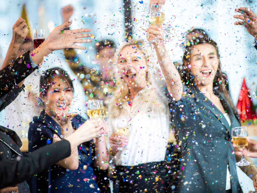 3 office workers raise a glass of wine at an office party, as confetti rains down on them.
