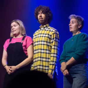 Three improv performers standing side by side on stage under blue lighting.