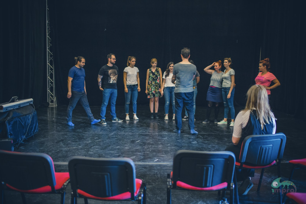 nine poeple stand upon a black stage in a semi circle, one stands in the middle with their back to us. @ chairs with backs to us are in the foreground.